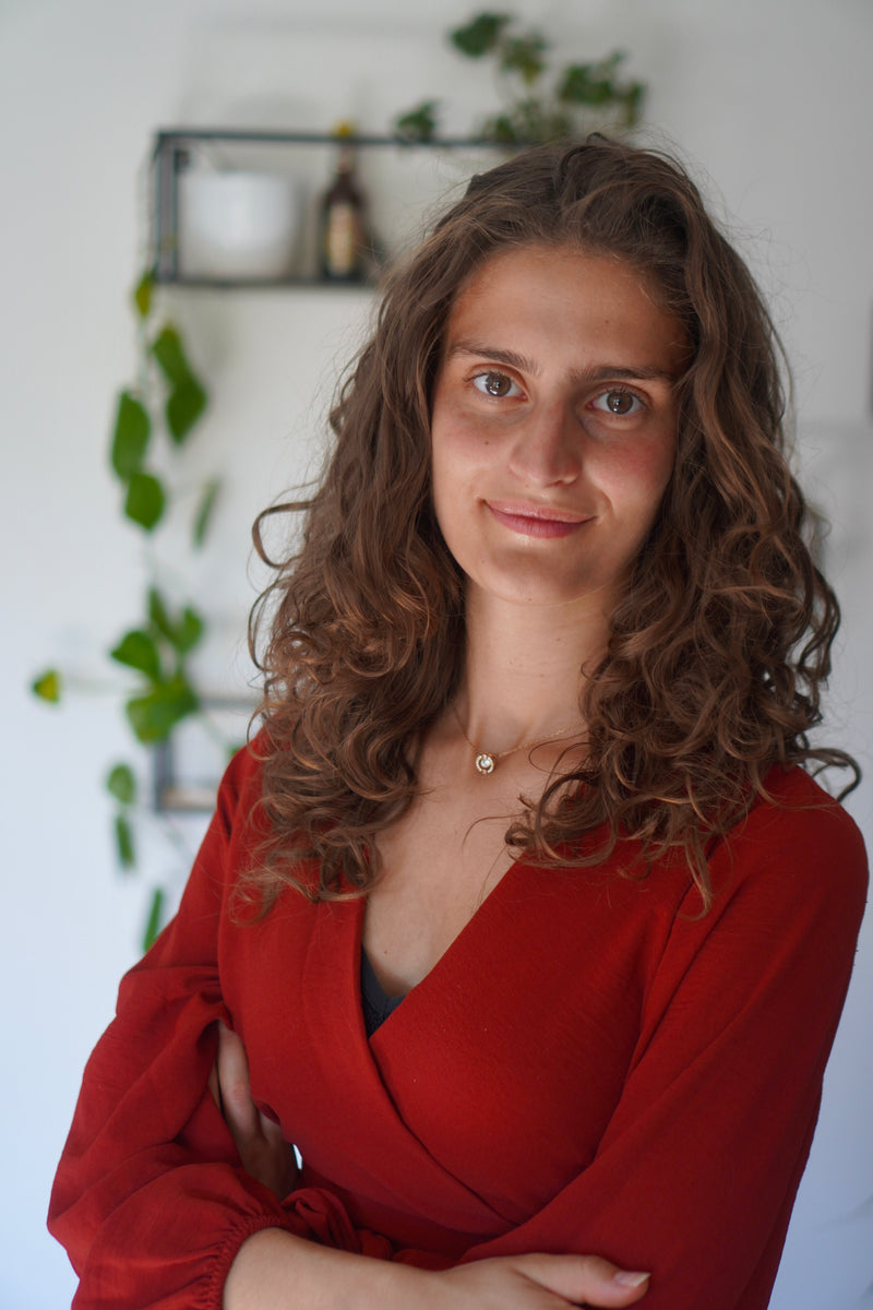 Person with curly brown hair wearing a red blouse, standing indoors with plants in the background.