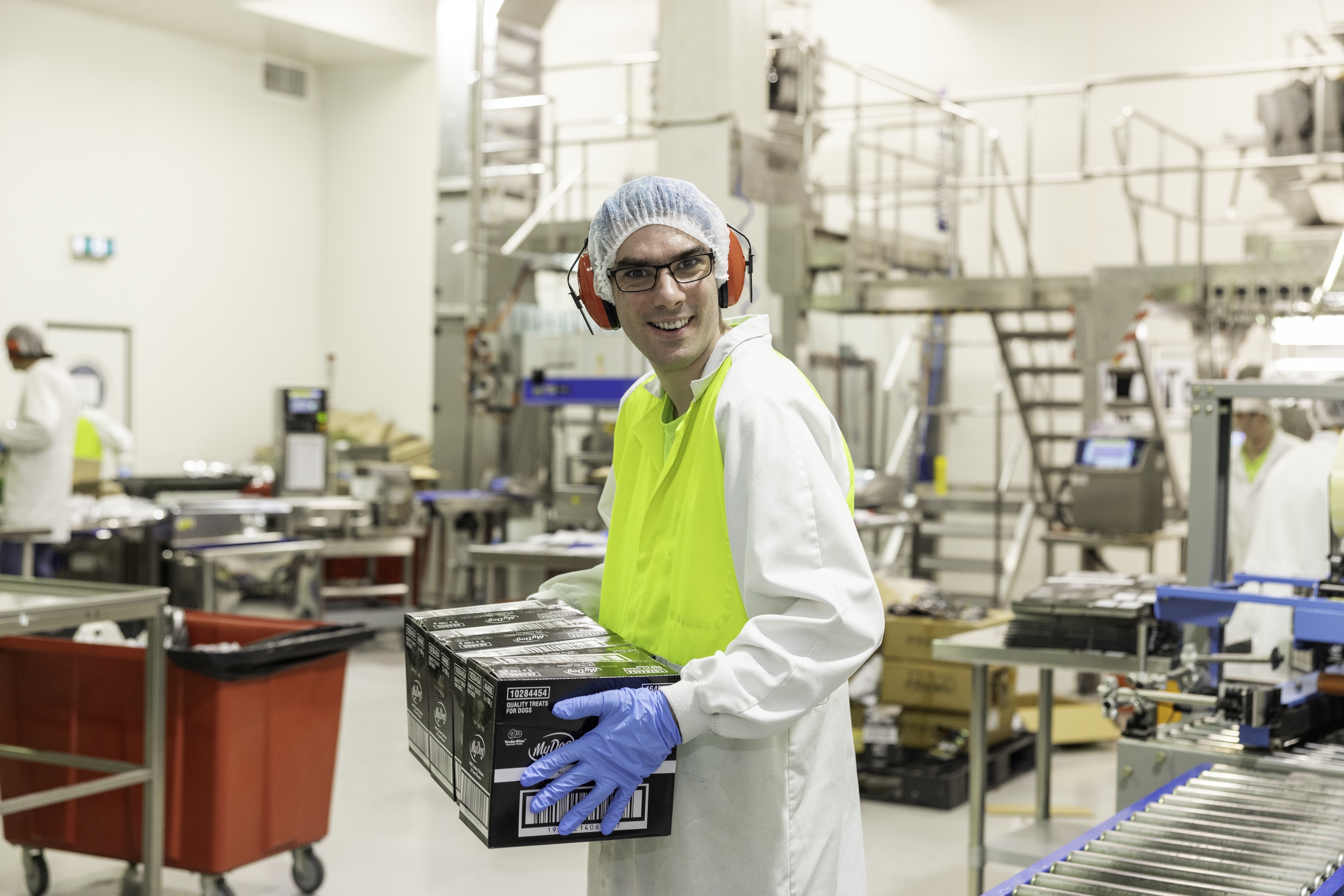 Factory worker in safety gear smiling while carrying packaged products on a production line.