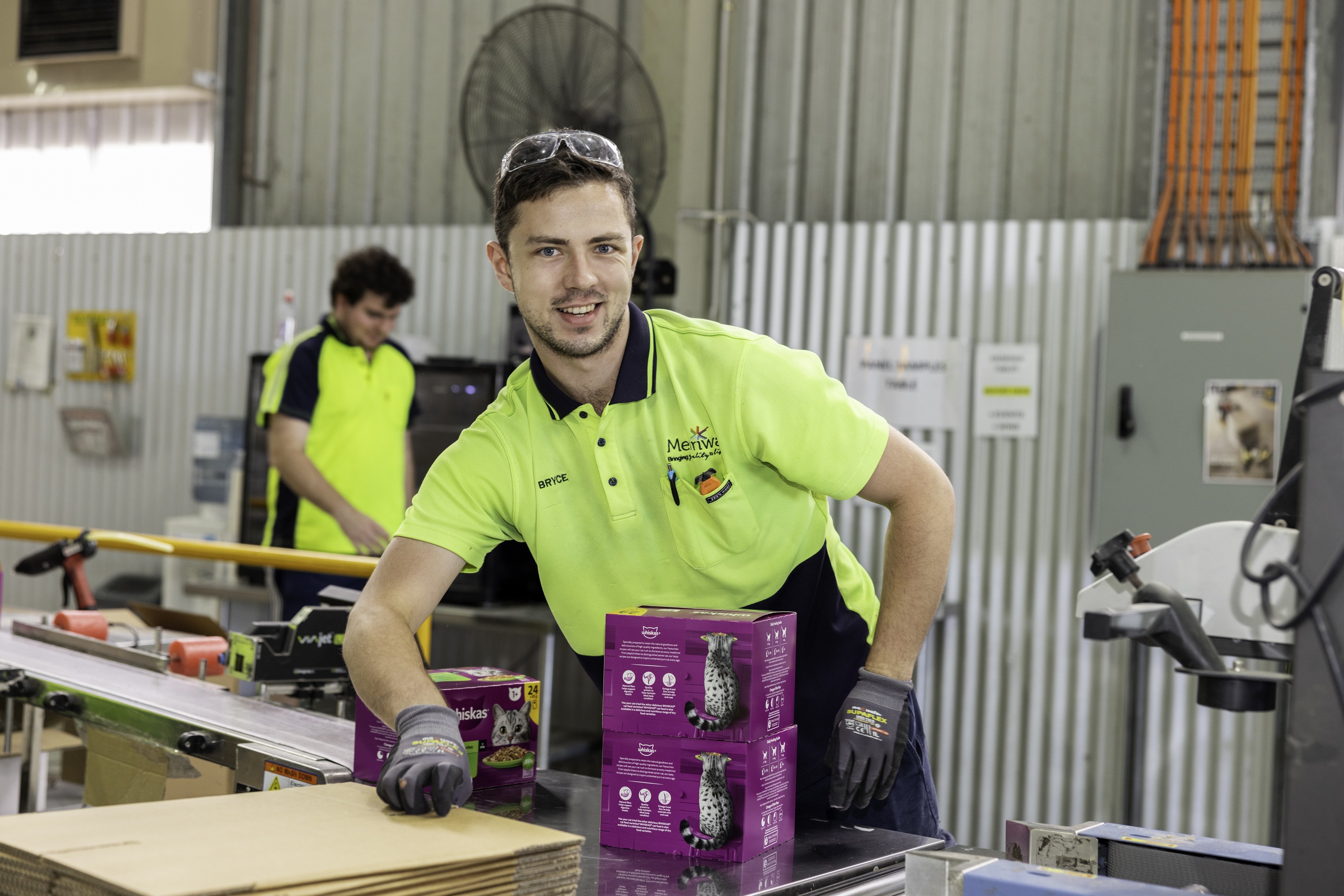 Worker in a hi-vis shirt smiles while packing pet food boxes on a production line in a warehouse.