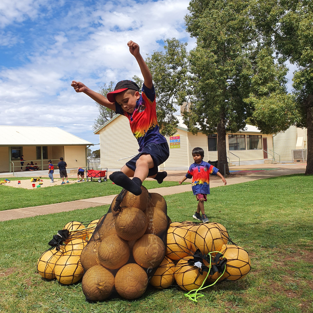 A child leaps off a stacked pile of netted yellow play balls on a school lawn while other children play nearby.