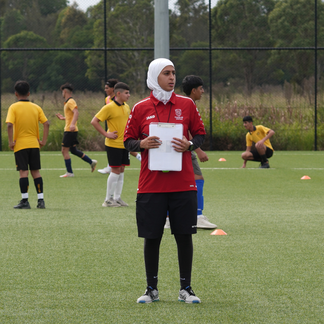 A coach in a red sports uniform and white hijab stands holding a clipboard on a soccer field while youth players in yellow shirts train in the background.