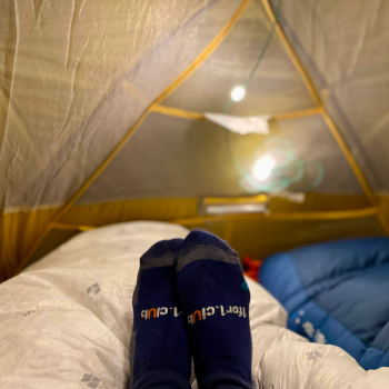 Feet in navy 1for1.club branded socks stretched out inside a glowing tent at night