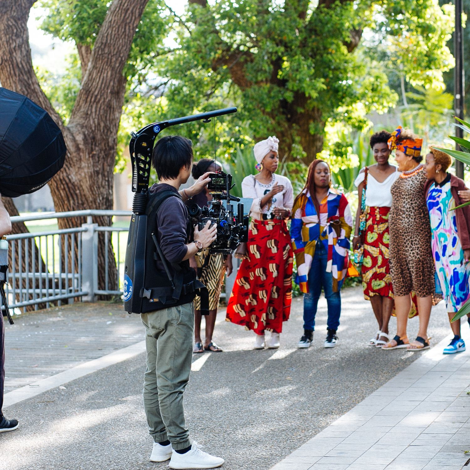 A filmmaker captures footage of a group of women dressed in colourful patterned outfits, standing together outdoors and engaged in conversation beneath large trees