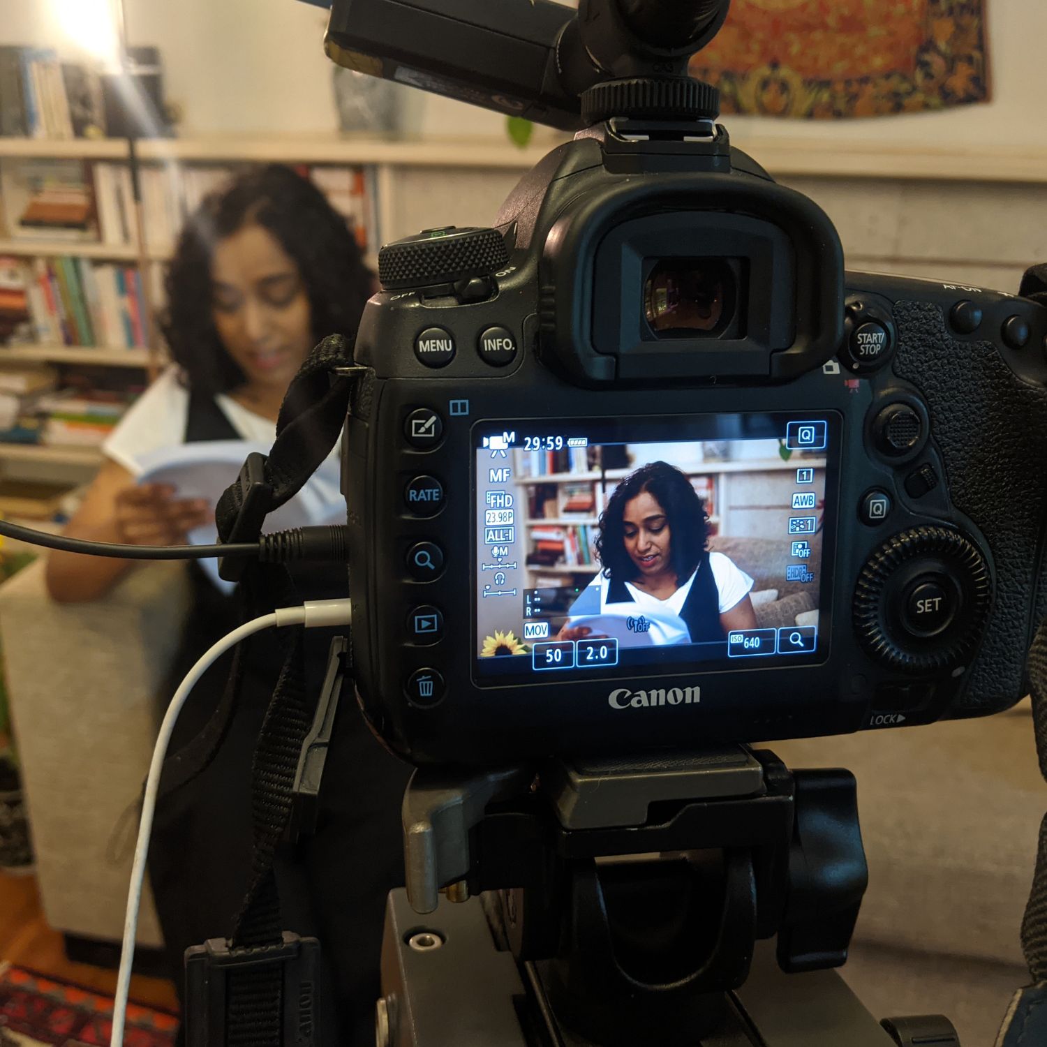 A camera captures footage of a woman sitting indoors reading from a book, with the camera screen showing her framed against a backdrop of bookshelves