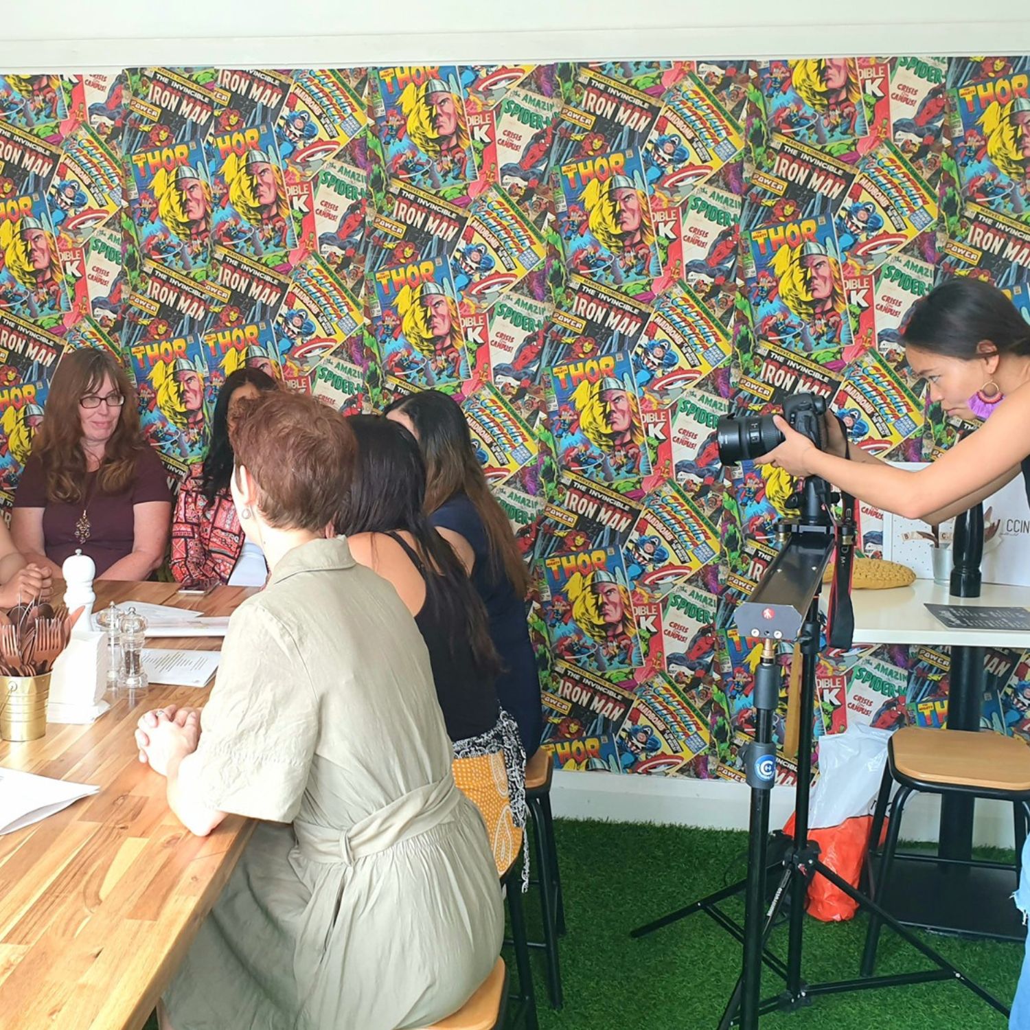 A photographer captures a group of women seated around a wooden table in front of a colourful comic book-themed wall, filming their discussion in a casual, creative setting