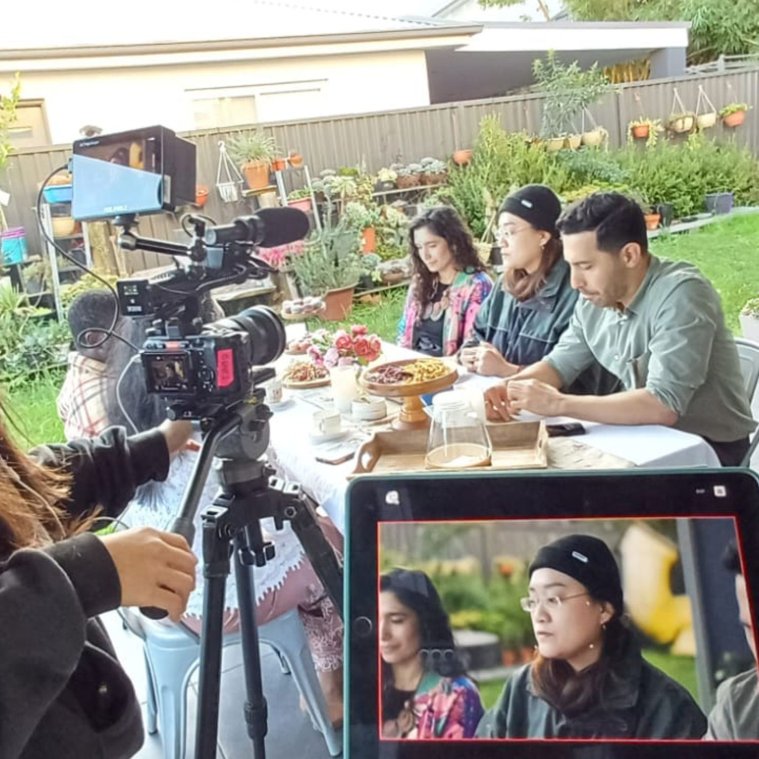 A camera crew films a group of people seated outdoors around a table set with food and drinks, capturing a discussion in a relaxed garden setting