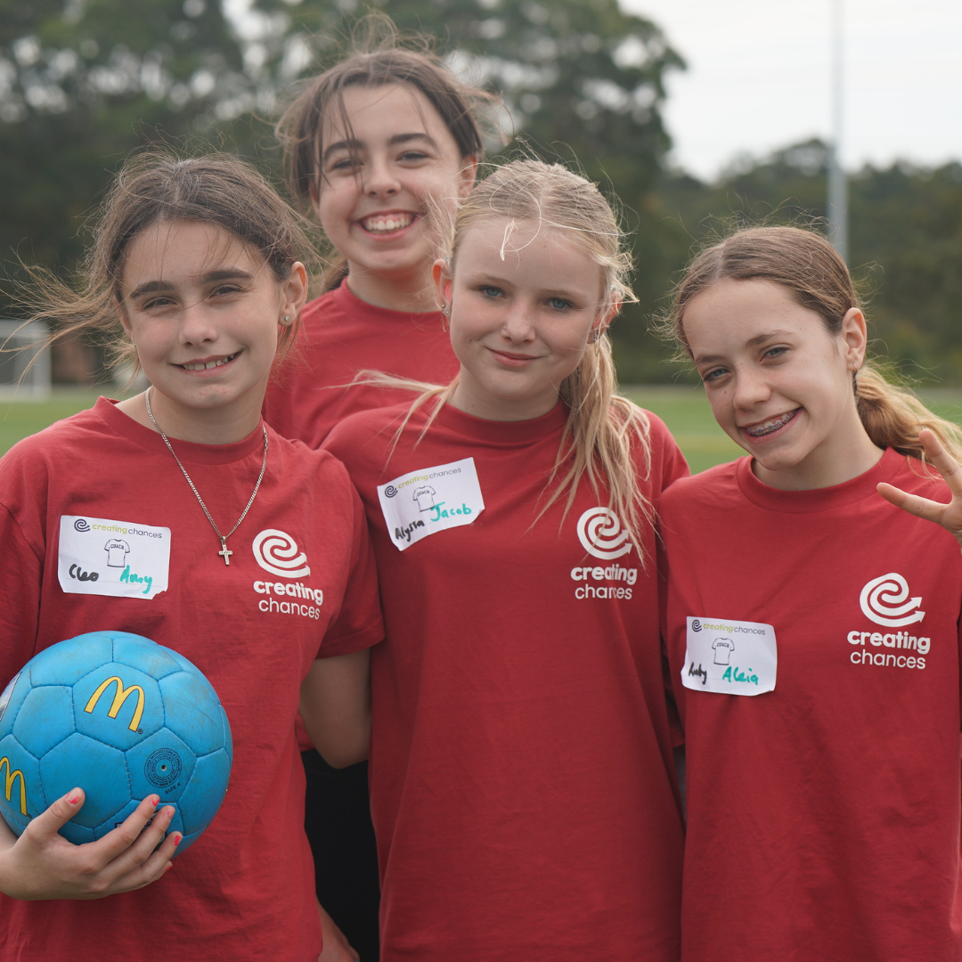 Four girls wearing red Creating Chances shirts smile together outdoors, holding a blue soccer ball.