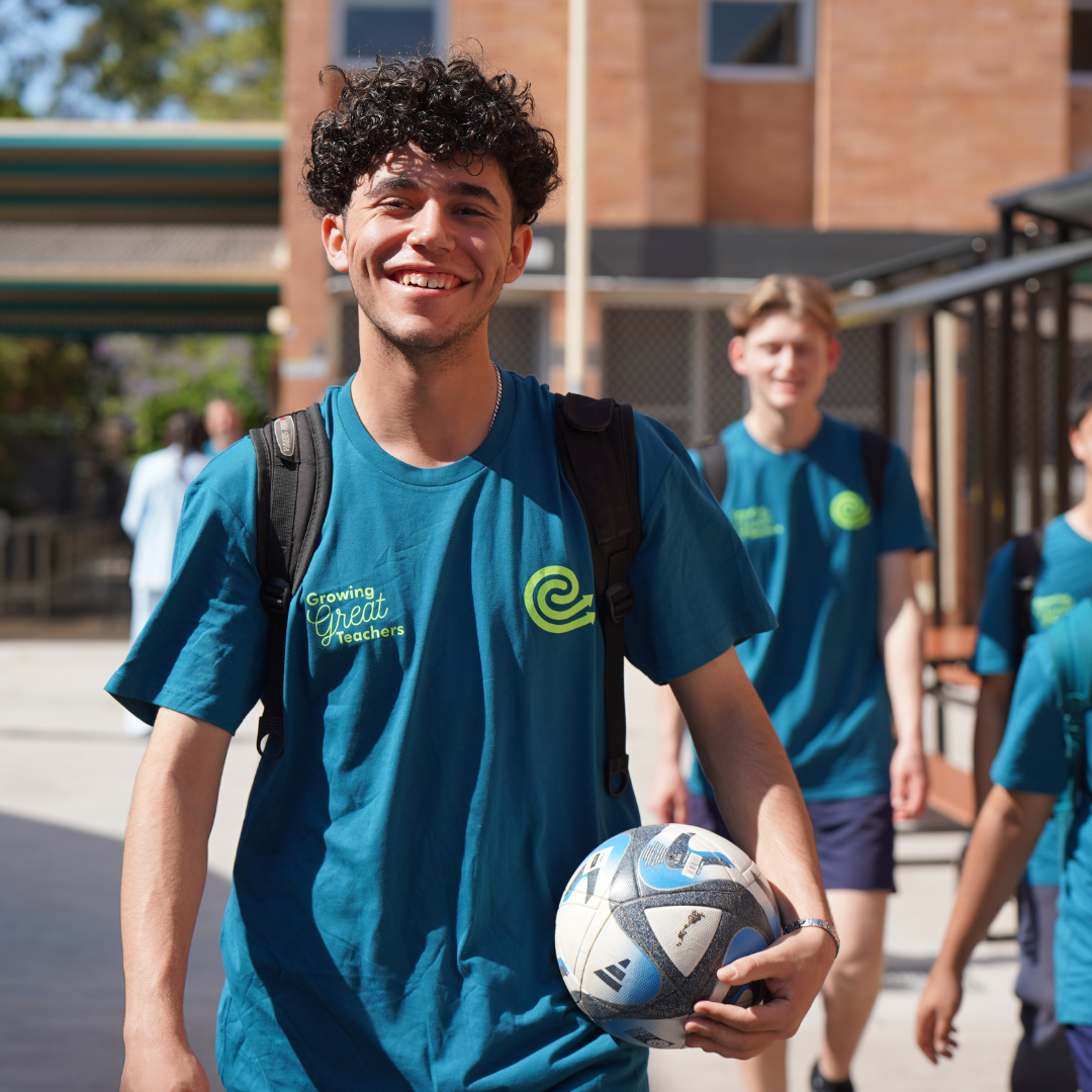 Young man smiling and walking through a school courtyard holding a soccer ball, with classmates following behind.