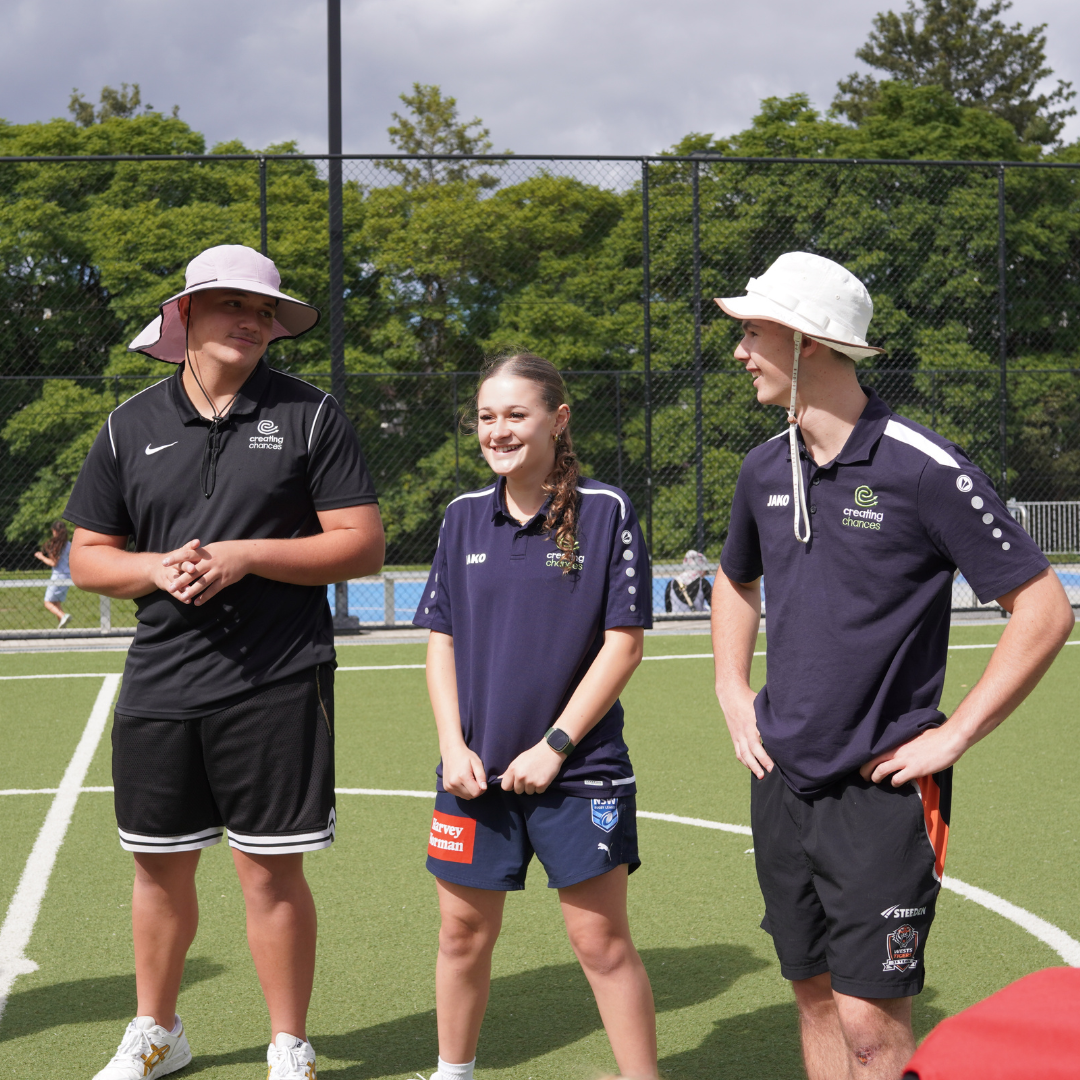 Three young coaches stand on an outdoor sports field, smiling and listening attentively during a youth training session.
