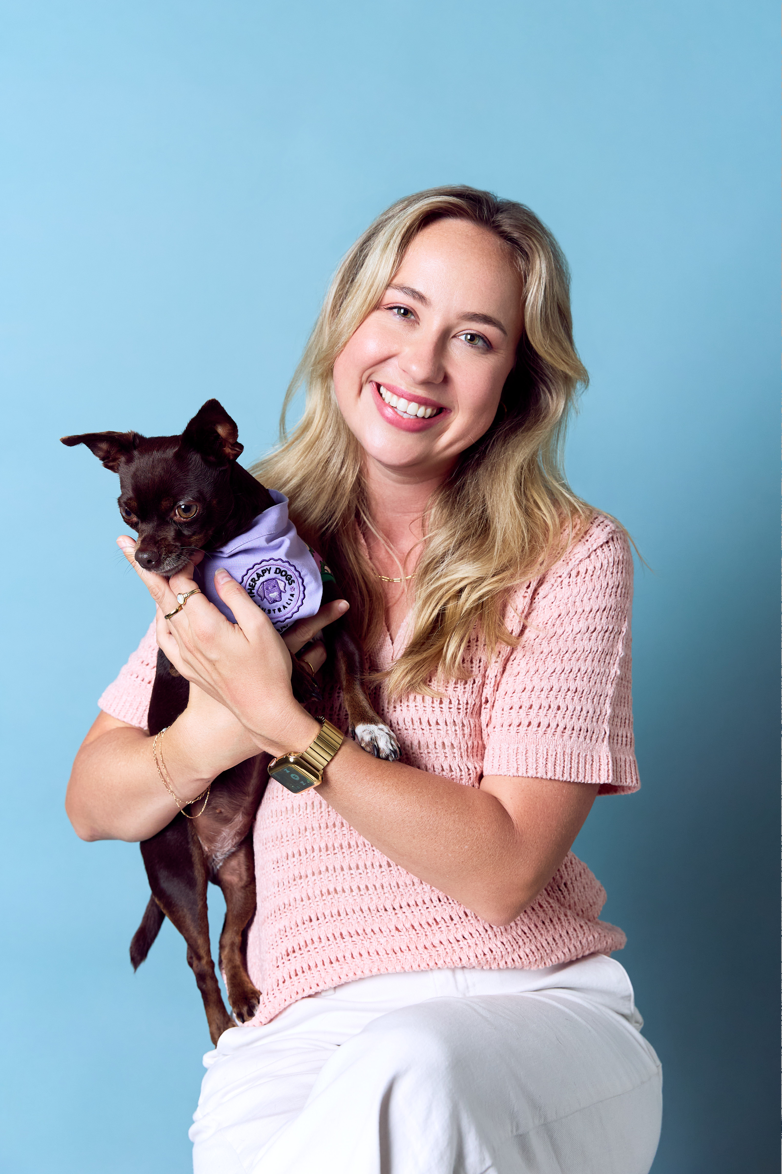 Smiling woman in a pink top holds a small brown dog wearing a purple vest against a blue background.