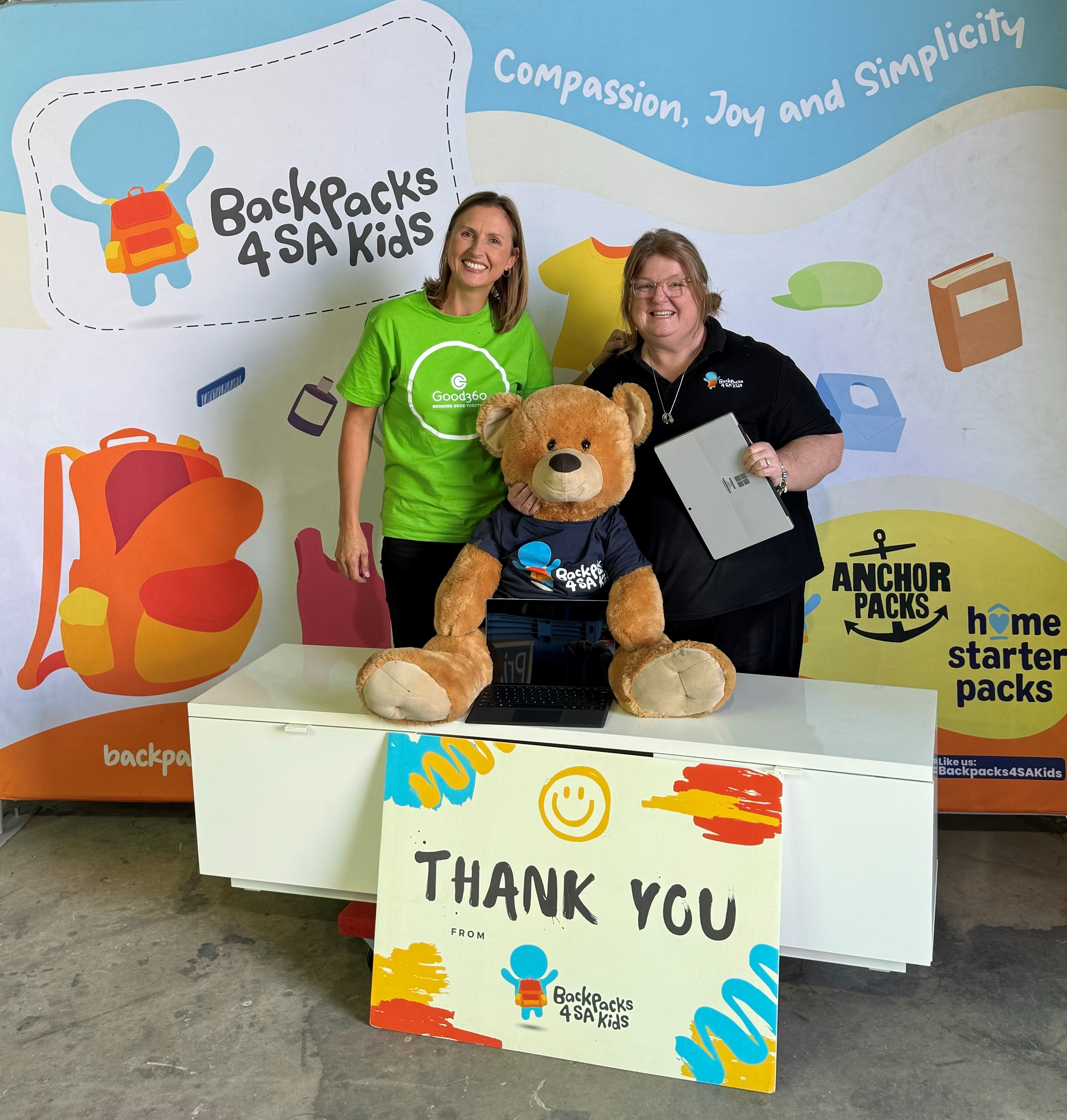 Two women stand beside a large teddy bear and a colourful ‘Thank You’ sign at a Backpacks 4 SA Kids display.