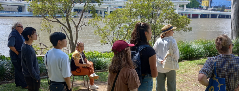 A group of people gathered by a riverside in a city park, listening to a seated speaker with buildings and a bridge in the background.