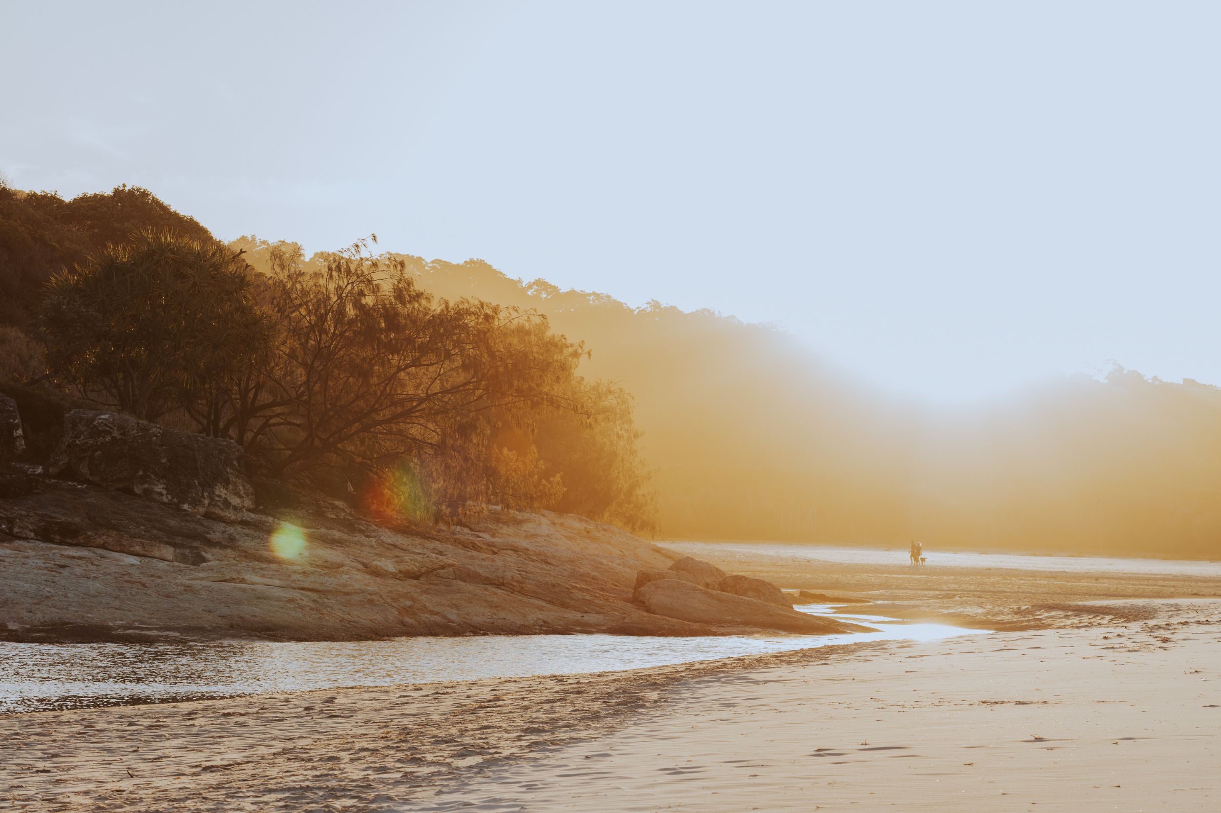 A peaceful beach scene with soft golden light illuminating trees and rocky hills in the background. The sandy shore and calm water reflect the sunlight, with two distant figures walking along the beach, creating a serene coastal atmosphere.