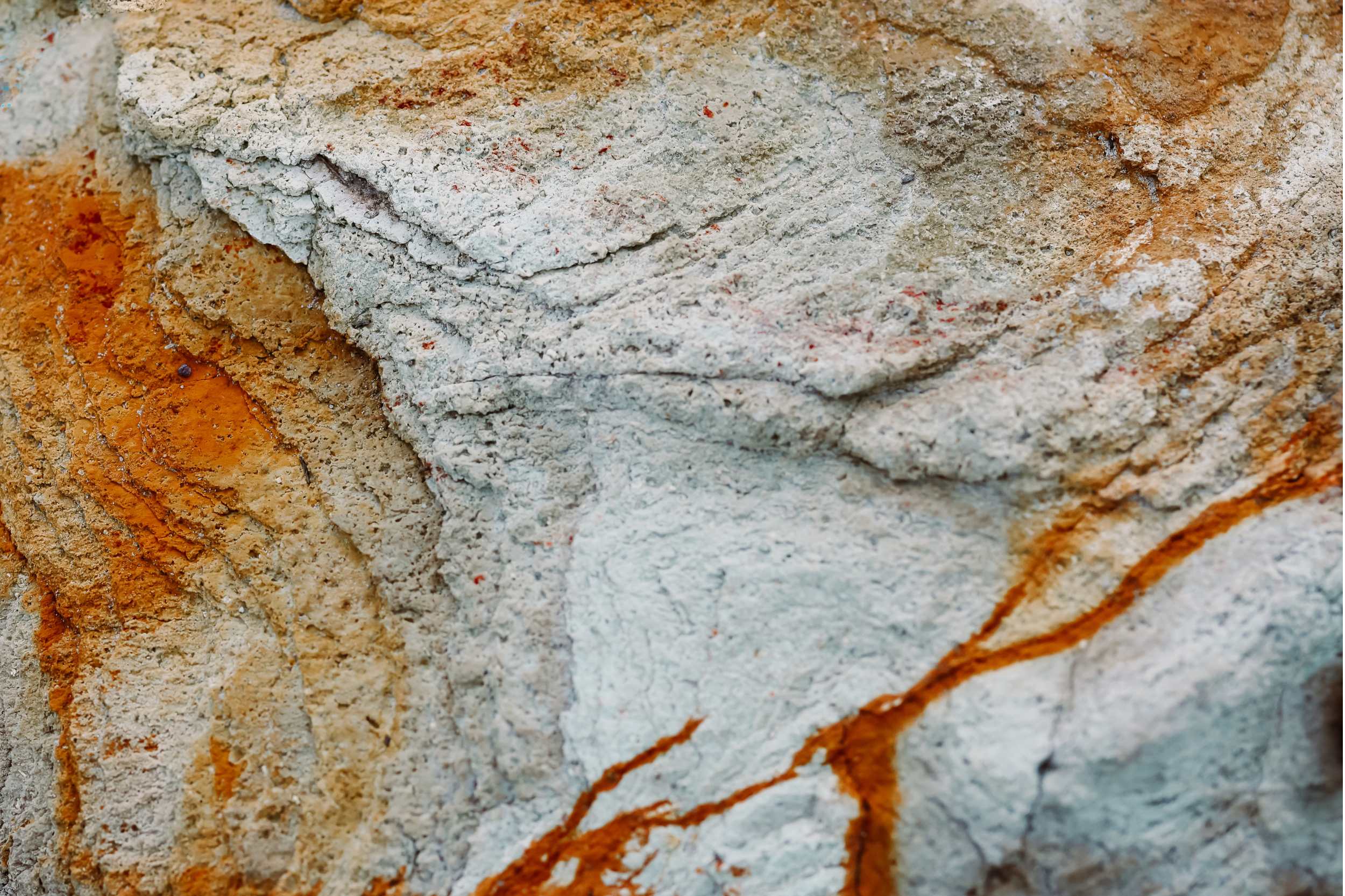 A close-up of a textured rock surface with layers of white, grey, and rusty orange tones, highlighting natural patterns and earthy colours.