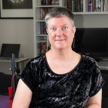 Professional headshot of a woman with short grey hair and gold hoop earrings seated in front of a home bookshelf