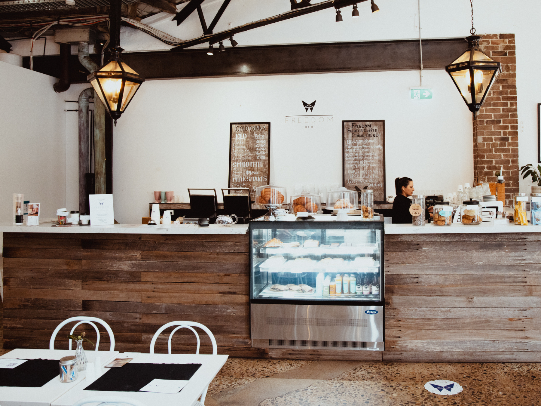 Image of The Freedom Hub (TFH; a social enterprise): A cozy cafe with a rustic wooden counter displaying pastries and snacks. A barista prepares orders behind a glass beverage case. Industrial-style lights hang overhead. Two white tables with menus and condiments are in the foreground. The "FREEDOM HUB" logo and menu board are on the wall.