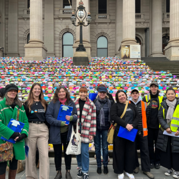 Group of people standing in front of Parliament House steps covered with colourful knitted squares arranged as part of a public display or community installation.