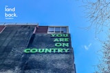 A building facade with neon green text reading "YOU ARE ON COUNTRY" against a blue sky, with "the land back. foundation" logo in the corner.
