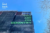 A building facade with neon green text reading "YOU ARE ON COUNTRY" against a blue sky, with "the land back. foundation" logo in the corner.