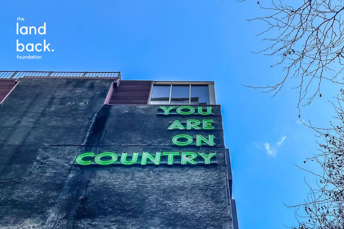 A tall, dark grey concrete building with a green neon sign that reads "YOU ARE ON COUNTRY." The sign is positioned on the upper part of the building, with the sky and some tree branches visible in the background. 