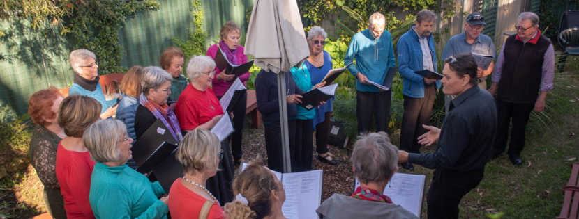 A choir of older adults stands in a garden, singing from sheet music while a conductor leads the group in an outdoor performance or rehearsal.