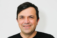 Headshot of a man with dark hair smiling in a black t-shirt against a plain white background.