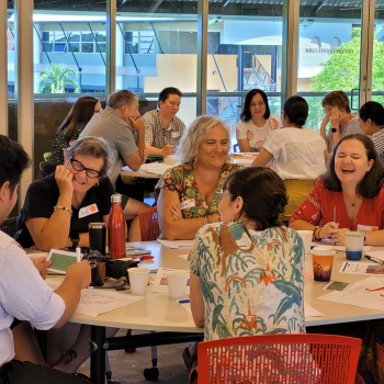Group of people laughing and talking around tables during a lively workshop session in a sunlit room.