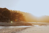 Golden sunlight illuminating a serene beach scene with soft sand, gentle waves, and distant trees on a rocky hill. A couple walks with their dog along the shoreline, surrounded by a warm, misty glow