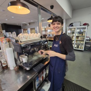 Woman in a navy apron smiling behind an espresso machine in a café.
