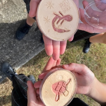 Two hands each holding a sand-filled disc etched with Aboriginal petroglyph designs including a turtle and a star motif.