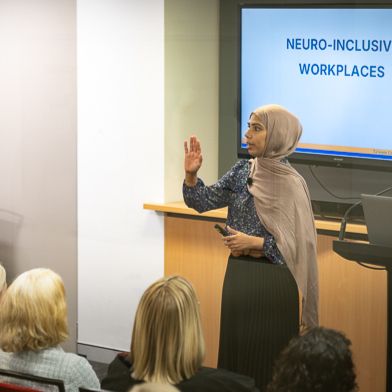 A woman in a headscarf presents to an audience in front of a screen displaying the words “Neuro-Inclusive Workplaces.”