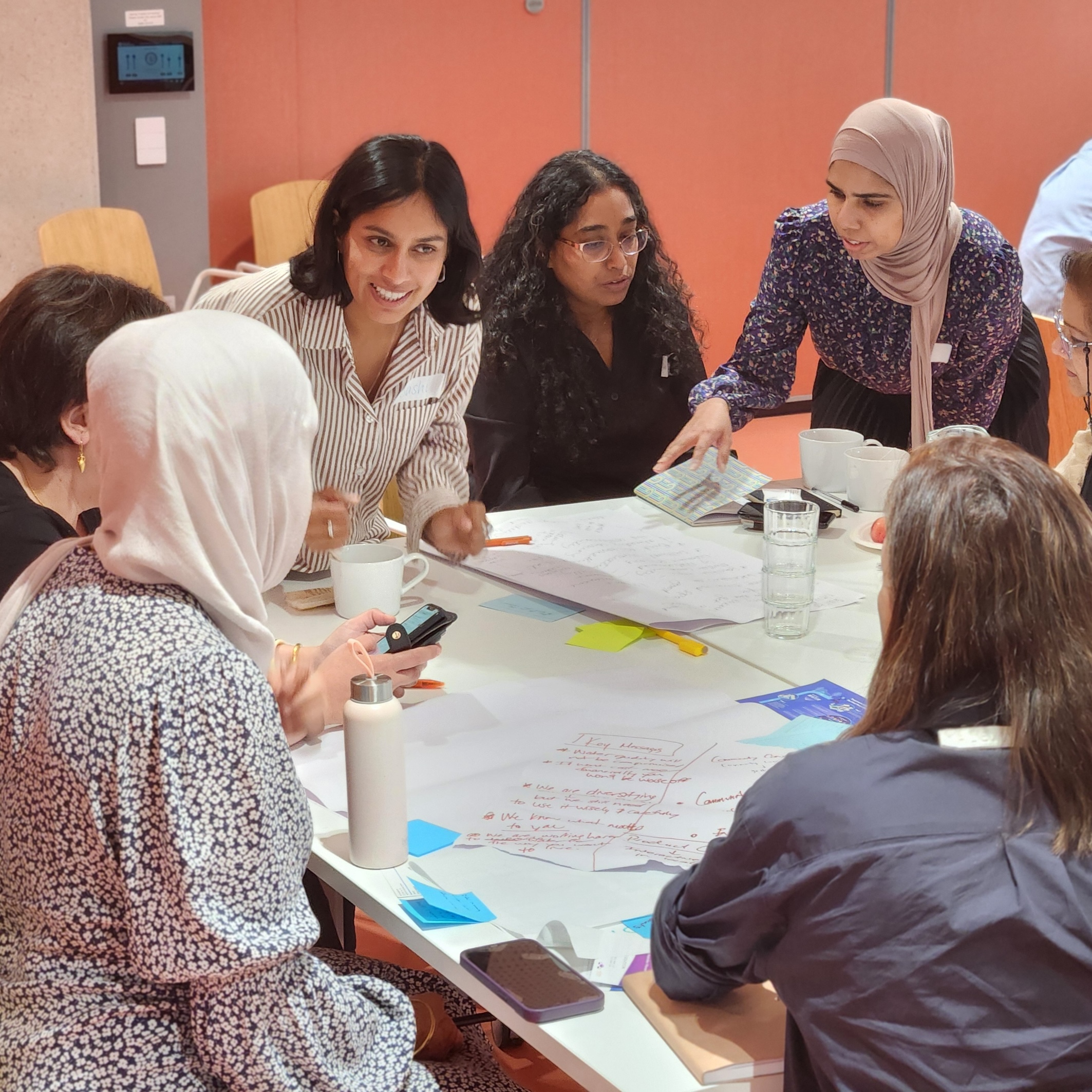 A diverse group of people collaborate around a table covered with notes, papers, and post-its during an interactive workshop session.
