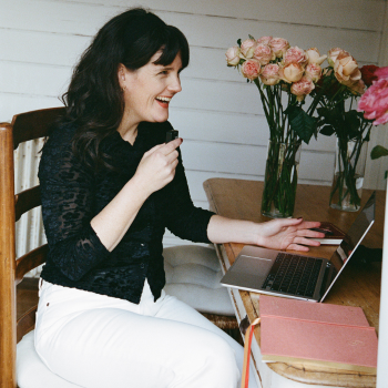 Woman laughing at a laptop on a wooden table set with vases of pink roses on a verandah.