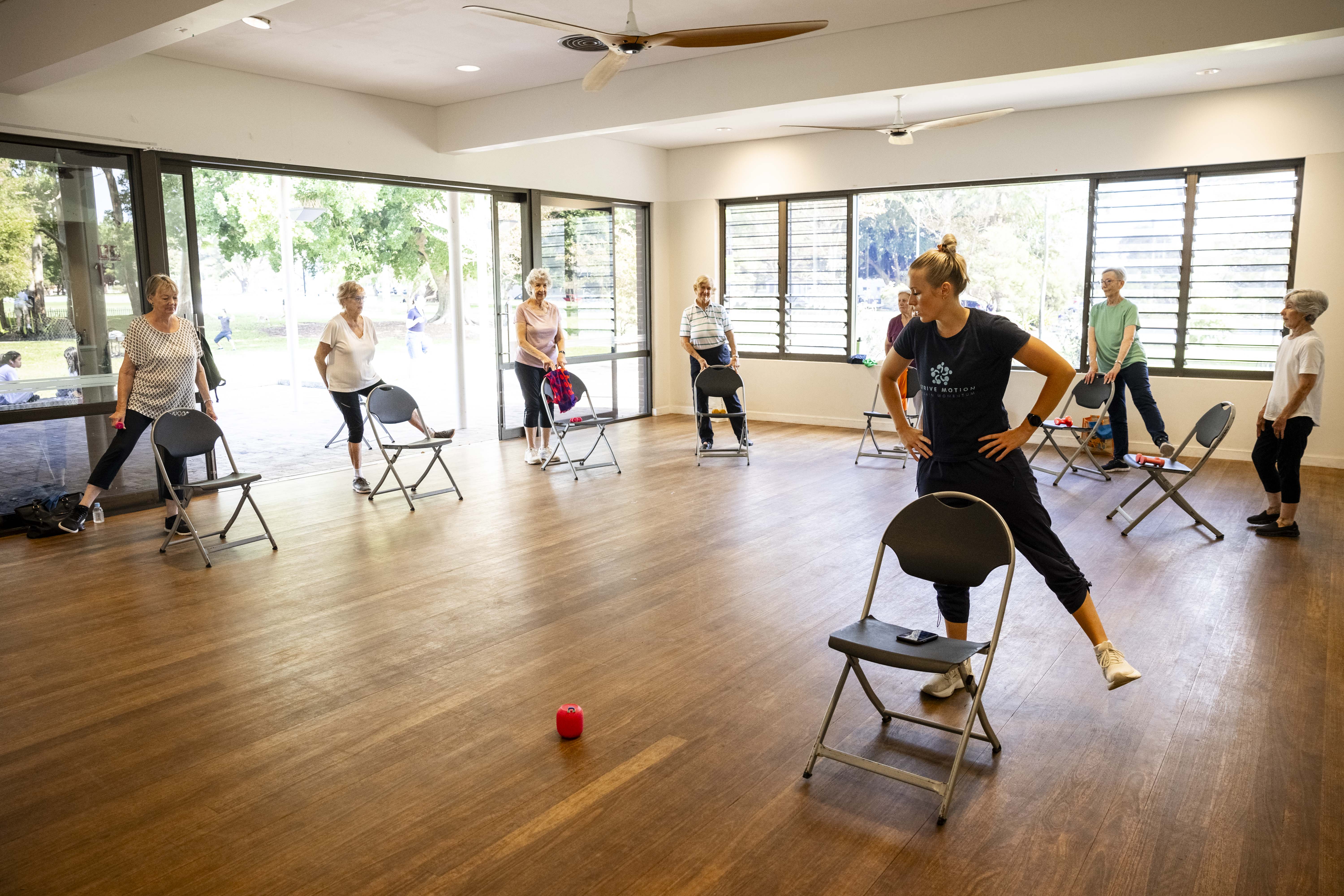 Instructor leads a seated exercise class with older adults using chairs in a community hall.