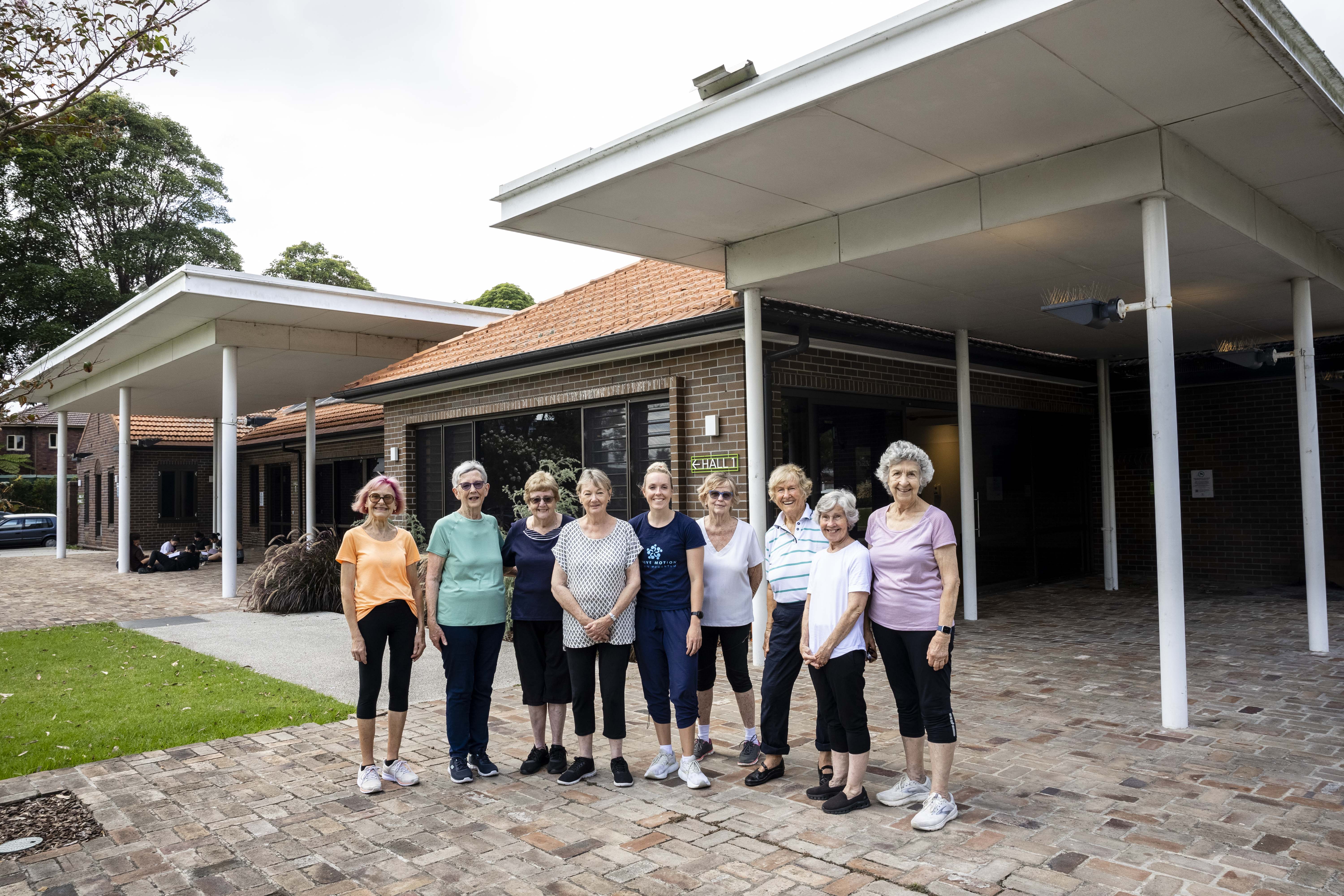 Group of older adults and an instructor stand smiling outside a community hall after an exercise session.