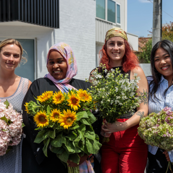 Four women standing outdoors, each holding a bouquet of flowers including sunflowers and pale blossoms, smiling together.
