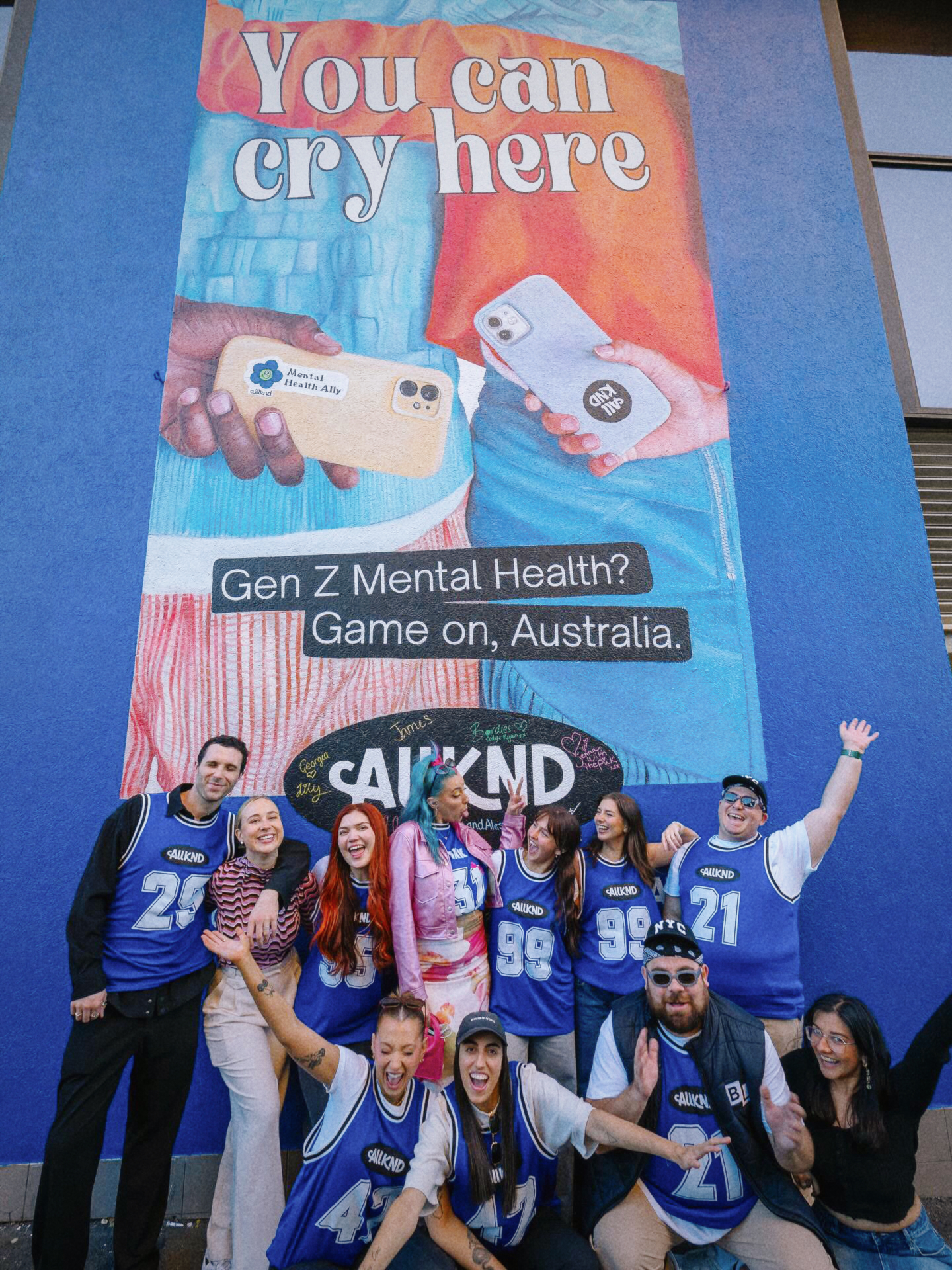 Group of young people in blue jerseys pose happily in front of a mural reading “You can cry here” promoting Gen Z mental health.