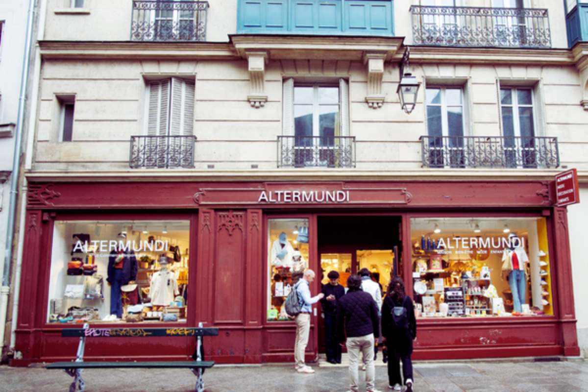 Storefront of "Altermundi" boutique with a burgundy facade and clothing displays in the windows, set beneath a heritage building with balconies, and people gathered near the entrance.