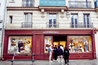 Storefront of "Altermundi" boutique with a burgundy facade and clothing displays in the windows, set beneath a heritage building with balconies, and people gathered near the entrance.
