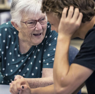 An older woman laughing warmly with a young volunteer leaning in close during a one-on-one digital support session