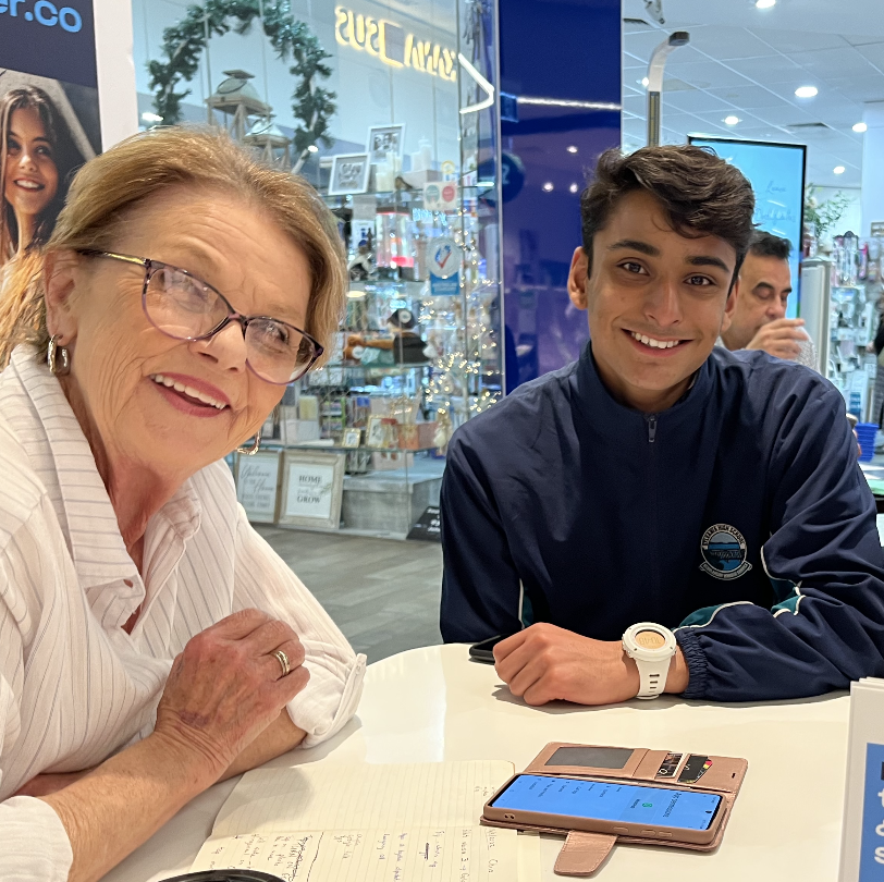 An older woman and a young Youngster.co volunteer smiling together at a table with a smartphone between them in a shopping centre setting