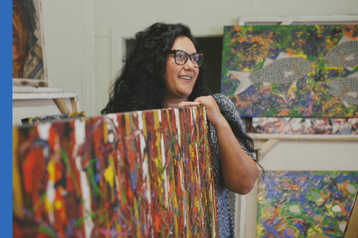 Smiling woman holding abstract artwork in a studio with other paintings.