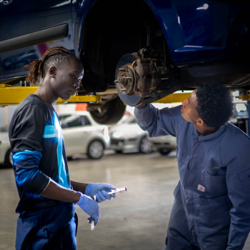 A young person holding a tool and watching attentively as a mechanic in overalls inspects a brake disc on a car raised on a hoist