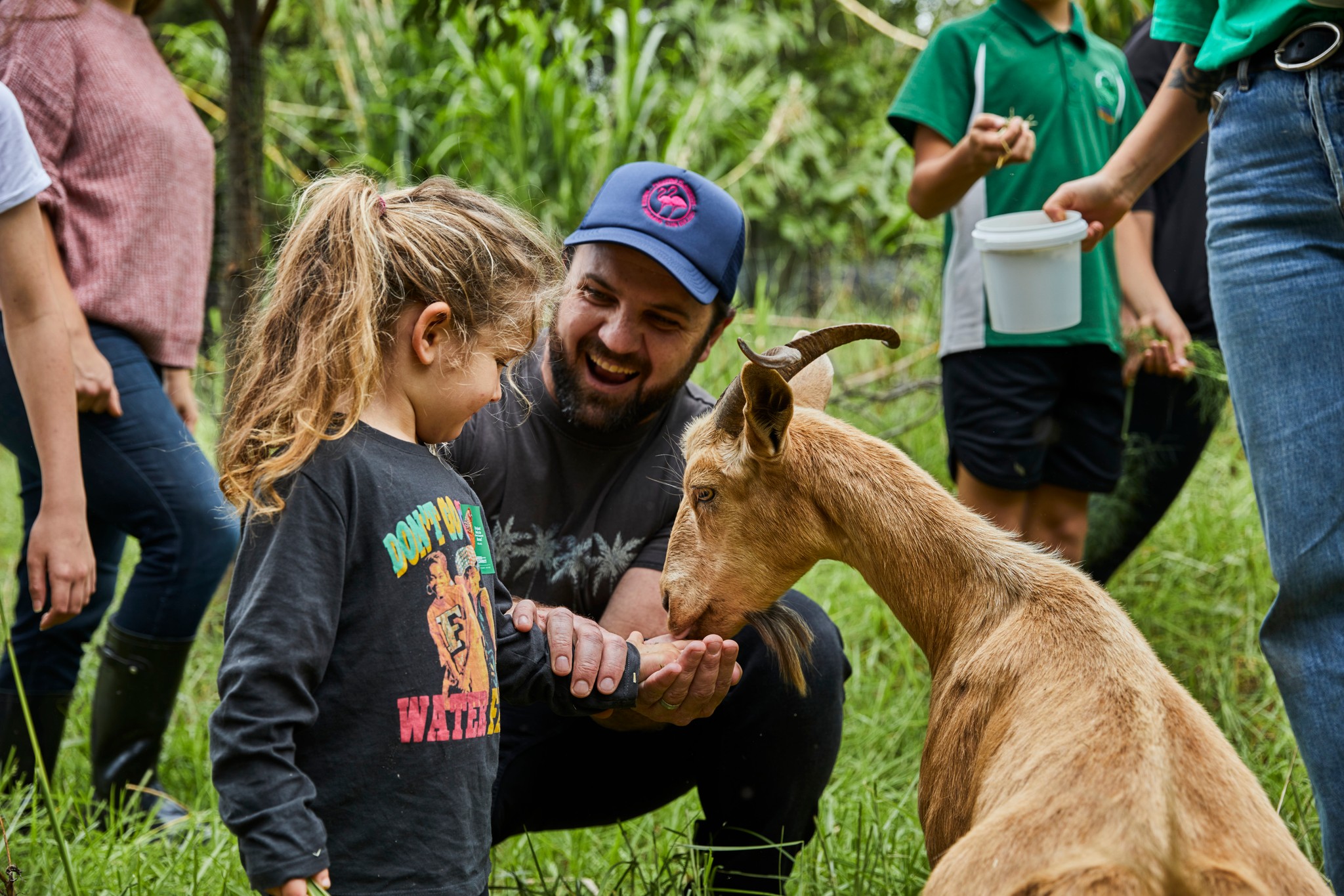 From Green Connect Farm, a smiling man helps a young girl feed a brown goat, surrounded by other visitors and lush greenery.